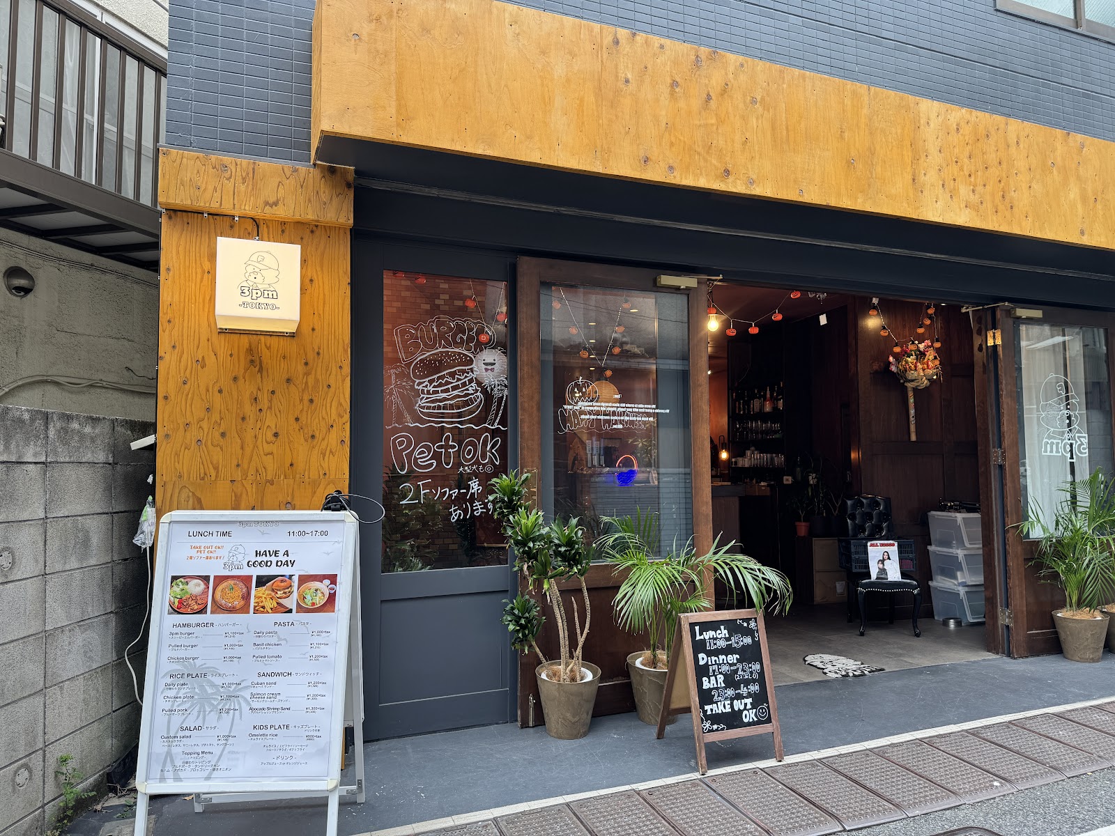 通り沿いの店先。合板の庇と壁、ガラスにチョークで描かれたバーガーの絵、店内にオレンジ色の豆電球、歩道に鉢植えのヤシ。