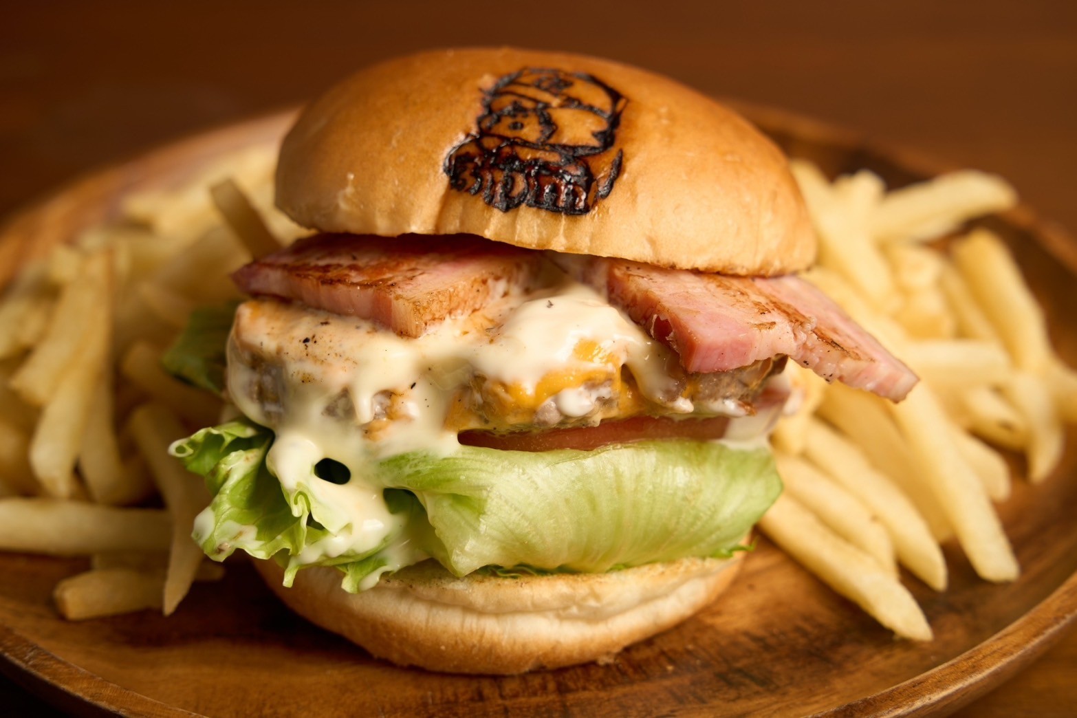 木の皿にのせたベーコンチーズバーガーと、まわりに積まれたフライドポテト。バンズには焼き印。